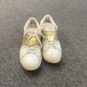 Valentino Garavani sneakers vit/guld - Säljer ett par snygga Valentino Garavani sneakers i vitt och guld. Skorna har klassisk snörning, platt sula och coola nitar bak på hälen. Tillverkade i skinn med gulddetaljer på sidan och ovandelen. Har inte kvar kartongen eller kvittot och sulorna är väldigt utnötta. Skriv om ni vill ha mer bilder 