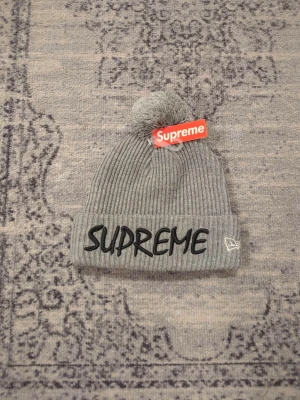 Supreme x New Era - Ribbstickad grå mössa från Supreme x New Era med stor bolltofs på toppen och uppvikt kant. Framtill står SUPREME broderat i svart och på sidan finns New Era-loggan. Klassisk streetwear-vibe och perfekt för kalla dagar.