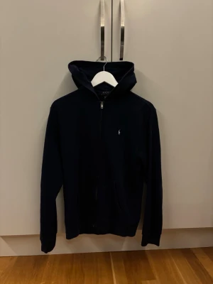 Polo Ralph Lauren Zip Hoodie strl 18-20 - En fin Polo Ralph Lauren hoodie med zip i en marinblå färg. Den är i Polo Ralph Laurens egna storlekar 18-20 vilket passar personer mellan 165-175cm långa. Skriv gärna vid frågor och funderingar✅