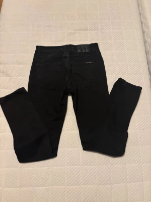 Svarta jeans från Nudie Jeans Co - Säljer ett par svarta jeans från Nudie Jeans Co med klassisk femficksdesign och diskret logga på bakfickan. Jeansen har en smal passform och är tillverkade i mjukt bomullsmaterial med stretch. Perfekta för dig som gillar stilrena och bekväma jeans.