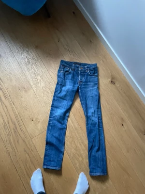 Blå Grim trim Nudie Jeans slim fit/straight fit - Säljer ett par klassiska blå jeans från Nudie Jeans med skinny fit. Jeansen har orangea sömmar, fem fickor och Nudie-loggan på bakfickan. Tillverkade i slitstark denim av bomull och har låg till normal midja. Perfekta för dig som gillar en smal siluett.