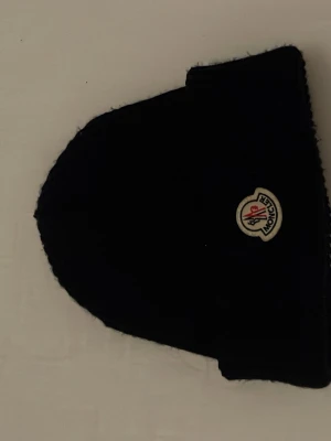 Mörkblå  Moncler mössa i ull - Säljer en svart Moncler mössa med uppvikt kant och klassisk logga framtill. Mössan är ribbstickad och gjord i mjuk ull som håller dig varm under kalla dagar. Perfekt accessoar för vintern och enkel att matcha med olika outfits.