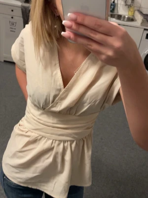 Beige omlottopp shein - En beige omlottopp från shein i storlek S. Toppen har korta ärmar, markerad midja, sitter åt lite baktill och omlottdesign framtill. Använd fåtal gånger. 