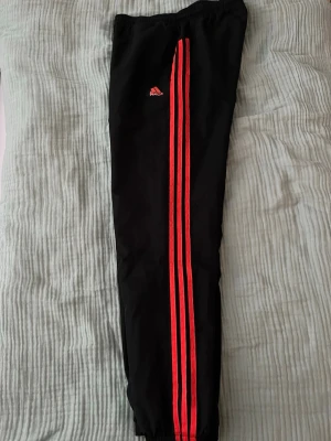 Svarta Adidas byxor med orangea ränder  - Säljer ett par svarta Adidas byxor med klassiska orangea ränder längs sidorna och Adidas-logga på låret. Byxorna har resår i midjan och vid bensluten, samt sidofickor. Tillverkade i lätt och glansig polyester, perfekta för träning eller chill. Storlek S men är mer som M skulle jag säga, finns även litet hål innersida högerben, se bild. 