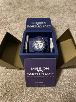 Omega x Swatch MoonSwatch Moonshine Gold Snoopy - Obs. Ej Original Säljer en Omega x Swatch MoonSwatch Mission to Earthphase Moonshine Gold Snoopy. Klockan har blå boett och armband, vit urtavla med blå och guldiga detaljer samt månfaser. Biokeramisk konstruktion och coolt tryck på baksidan. Perfekt för dig som gillar unika accessoarer.