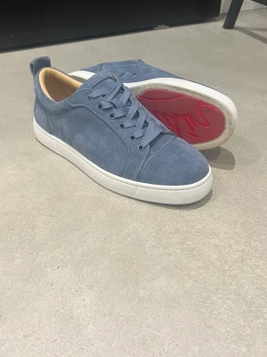 Blå mockasneakers från Christian Louboutin - Säljer ett par snygga blå sneakers från Christian Louboutin i mocka med klassisk röd sula och vita kanter. Skorna har rund tå, snörning och beige innersula med guldfärgad logga. Perfekta för dig som vill ha något exklusivt och stilrent.