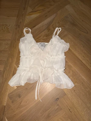 Vit volang topp med knytband - Supersöt vit linnetopp  med tunna axelband, volangdetaljer och knytband framtill. Toppen har v-ringning och är tillverkad i ett lätt, luftigt material som känns perfekt för varma dagar.