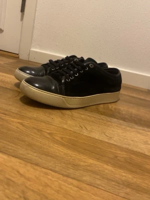 Svarta sneakers från Lanvin i skinn och mocka - Säljer ett par svarta Lanvin sneakers med ovandel i mocka och detaljer i blankt skinn på tån. Skorna har vita, tjocka gummisulor och klassisk snörning. Perfekta för dig som gillar stilrena och lyxiga sneakers med en clean vibe.