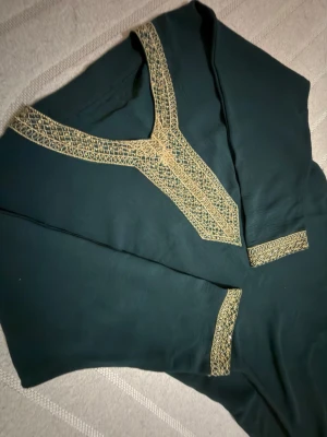Marockansk mörkgrön långklänning med guldbrodyr - Säljer en elegant mörkgrön långklänning med detaljerad guldbrodyr runt halsringningen och ärmsluten. Klänningen har trekvartsärm och en mjuk, följsam passform. Materialet känns lätt och svalt, perfekt för speciella tillfällen. Klänningen passar alla som mellan storleken S till L alltså om man har storleken S så blir det baggy vilket ska vara samma sak med L fast mindre oversized. Är öppen för att diskutera pris