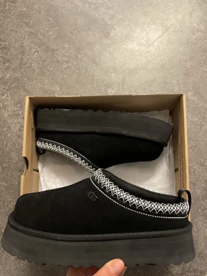 Svarta UGG slip-ons med mönstrad kant - Svarta slip-on skor från UGG med fluffigt foder och grov sula. Skorna har en snygg vit och svart mönstrad kant runt öppningen och är tillverkade i mocka. Perfekta för kalla dagar när du vill vara både bekväm och stilren.