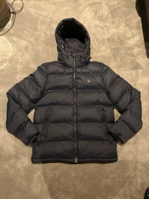 Mörkblå pufferjacka från GANT - Snygg mörkblå pufferjacka från GANT med huva och dragkedja framtill. Jackan har två sidofickor med dragkedja och GANT-logga på bröstet. Perfekt för kalla dagar och riktigt skön att bära. Klassisk och stilren design.