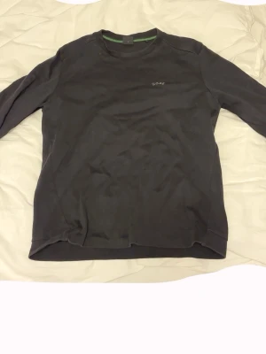 Svart långärmad tröja från BOSS - En stilren svart långärmad tröja från BOSS med diskret logga på bröstet. Klassisk rund halsringning och mjukt material som känns skönt mot huden. Perfekt till jeans eller chinos för en clean look.