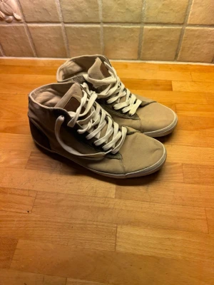 Beige höga sneakers med vita snören - Säljer ett par beige höga sneakers med vita snören och bruna detaljer på hälen. Skorna har en klassisk rund tå och platt sula. Ovandelen är i tyg och sulan är i gummi. Perfekta för dig som gillar enkel och clean stil använda två gånger.
