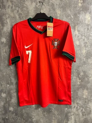 Fotbollströja Portugal Ronaldo #7 - Large - Portugal fotbollströja från Nike med nummer 7 och Ronaldo på ryggen. Tröjan är röd med gröna och svarta detaljer vid krage och ärmslut, samt broderad landslagssköld på bröstet. Perfekt för dig som vill stötta Portugal på match!
