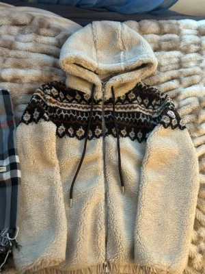 Moncler fair isle - Säljer en beige teddyjacka från Moncler med huva och dragkedja. Jackan har ett snyggt brunt och svart mönster över bröstet och axlarna samt långa snören i huvan. Mysigt teddy-material som är perfekt för kyliga dagar. Moncler-logga på ärmen för extra stilpoäng. Storlek S men sitter som M så passar dig som är 175-190 