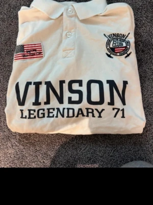Vit pikétröja Vinson Legendary 71 storlek L - Säljer en vit pikétröja från Vinson med trycket 'VINSON LEGENDARY 71' på bröstet. Tröjan har klassisk krage, två knappar, korta ärmar och detaljer som amerikansk flagga och broderad logga. Perfekt till en sportig och avslappnad stil.
