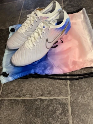 Vita Nike fotbollsskor med blå detaljer - Säljer ett par vita Nike fotbollsskor med snygga blå och guld detaljer på hälen. Skorna har snörning och en smidig, strömlinjeformad design med rund tå och platt sula. Perfekta för dig som vill sticka ut på planen. Ordernummrr får man till