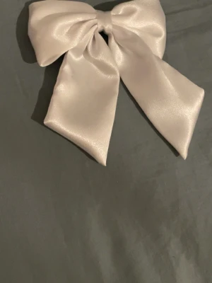 Stor ljusrosett i satin - Säljer en stor och lyxig rosett i ljus satin med hårspänne på baksidan. Perfekt accessoar för att piffa upp frisyren med en elegant touch. Rosetten har en mjuk glans och breda band som ger ett statement-utseende.