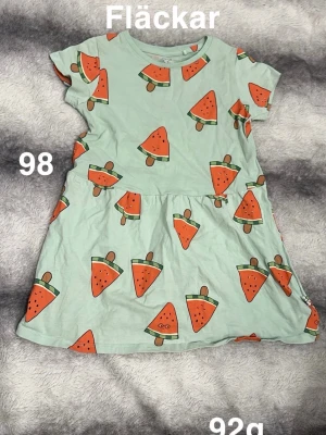 Mintgrön klänning med vattenmelonmönster - Supersöt mintgrön klänning med korta ärmar och ett lekfullt mönster av vattenmeloner över hela plagget. perfekt för varma dagar. Kjolen har en lätt utställd form som ger en skön och ledig känsla.🌸 Fläckar. Storlek 98
