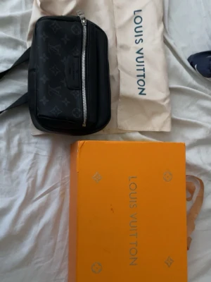 Svart axelväska  - Säljer en svart axelväska  med klassiskt monogrammönster och silverfärgad dragkedja. Väskan har en kompakt och fyrkantig form, perfekt för att bära det viktigaste. Kommer med dustbag och originalkartong.