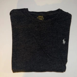 Polo Ralph Lauren Mörkgrå T-Shirt - Mörkgrå t-shirt från Polo Ralph Lauren i custom slim fit. Klassisk design med rund hals och den ikoniska broderade loggan på bröstet. Tillverkad i mjuk bomull som känns skön mot huden. Perfekt för en clean och stilren look.
