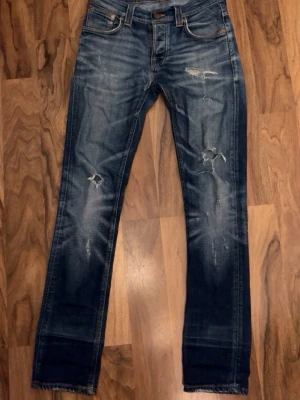 Nudie jeans  - Säljer ett par blå  jeans med slitningar och distressed detaljer på både fram- och baksida. Jeansen har klassisk femficksmodell, låg midja och är tillverkade i jeansmaterial. Perfekta för en avslappnad och trendig look. STORLEK 28/32. Finns ett litet hål med det synes knappt. 