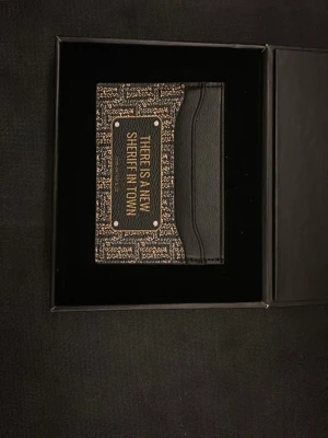 Plånbok John Hatter & Co svart mönstrad - HELT OANVÄND plånbok från John Hatter & Co i svart skinn med beige mönster och texten 'THERE IS A NEW SHERIFF IN TOWN' på. Fack på båda sidorna och ett mynt fack i mitten.                            Ny pris - 695kr 