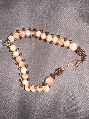 Handgjord pärlväst med ekollonberlock, namn”light.brown.white.kind.of.flower,silver.rose”   - Unik handgjord väst med pärlor i beige, brunt och vitt. Västen har små glaspärlor i brunt och större marmorerade pärlor, samt en silverfärgad ekollonberlock. Stängs med metallhake. Perfekt för dig som vill sticka ut med detaljer. Liknade som de förra Namn ”light.brown.white.kind.of.flower,silver.rose” artikelnummer ”064W” Stolen 16 cm pris 150kr