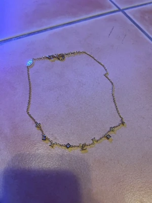 Halsband  - Stilrent och feminint halsband med dekorativa detaljer och gnistrande stenar. Perfekt för både vardag och fest – lika snyggt ensamt som tillsammans med andra smycken i lager-på-lager-stil.  🔹 Färg: Guldfärgad 🔹 Design: Tunt kedjehalsband med små symbol- och sten-detaljer 🔹 Skick: Mycket fint skick 🔹 Lås: Karbinlås 🔹 Passform: Bekvämt och lätt att bära  Ett trendigt smycke som lyfter vilken outfit som helst 