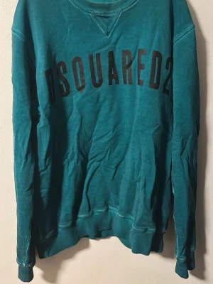 Grön sweatshirt från Dsquared2 - Säljer en grön sweatshirt från Dsquared2 med svart tryck över bröstet. Tröjan har rund halsringning, långa ärmar och ribbade muddar vid ärmslut och nederkant. Perfekt för dig som gillar streetwear och vill ha något bekvämt och snyggt. 