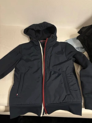 Mörkblå vindjacka från Tommy Hilfiger - Snygg mörkblå vindjacka från Tommy Hilfiger med huva och dragkedja i rött och vitt. Jackan har två fickor med dragkedja och diskret logga på ärmen. Materialet är polyester och jackan har en sportig vibe med klassiska Hilfiger-detaljer. Storlek xs men passar också S.