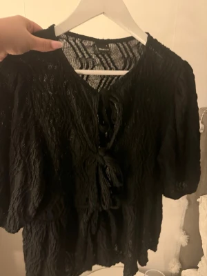 Svart blus med knyt och spetsdetaljer - Säljer en svart blus från Gina med transparent spets och snygga knyt framtill. Blusen har korta puffärmar och en luftig, vågig struktur i tyget som ger en cool look. Perfekt att styla med jeans eller kjol för en trendig outfit.