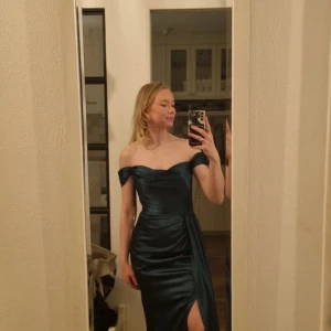 Mörkgrön axelbandslös klänning - Elegant mörkgrön axelbandslös klänning i satin med en smickrande slits framtill. Perfekt för en festlig kväll. Klänningen har en draperad design som ger en lyxig känsla. Fin figursydd med midja och höfter blir tydliga. Inbyggd bh. Medelhög rygg. Jag är 169cm och klänningen är perfekt lång med klackskor. Helt ny och oanvänd. Nypris är 4500kr. Pris går att diskutera💕😍