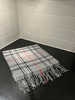 Grå rutig halsduk med fransar - Säljer en stilren grå halsduk med klassiskt rutigt mönster i svart, vitt och röda detaljer. Halsduken har fransar i ändarna och är mjuk och lätt att styla till olika outfits. Liknade burberry 
