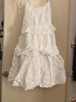 Vit volangklänning med knytband - Supersöt vit kortklänning med tunna axelband som knyts och volangdetaljer i flera lager. Klänningen har en luftig passform och är perfekt för varma dagar. Tillverkad i ett lätt bomullstyg som känns skönt mot huden. Dent justerbar i axlar och rygg.