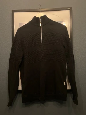 Svart stickad tröja med half zip - Säljer en svart stickad tröja med hög krage och half zip-dragkedja framtill. Tröjan har ett strukturerat mönster över bröstet och ärmarna samt långa ärmar. Perfekt för kyligare dagar och enkel att matcha med jeans eller byxor. Lite sliten i färgen!
