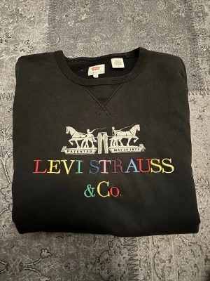 Svart sweatshirt från Levi's med brodyr - Säljer en svart sweatshirt från Levi's med färgglad broderad logga och klassiskt tryck framtill. Tröjan har rund halsringning och långa ärmar. Perfekt för dig som gillar streetwear och vill ha något bekvämt och stilrent. OBS! stor i storleken!
