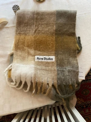 Rutig Acne Studios Scarf - Säljer en fluffig och mjuk scarf från Acne Studios i mohairmix med rutigt mönster i beige, grått, vitt och brunt. Scarfen har fransar nedtill och en stor Acne Studios-logga framtill. Perfekt för lager-på-lager och att ge din outfit en cool touch. Aldrig använd utomhus, endast testad inomhus!