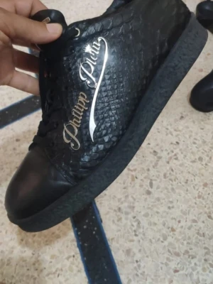 Svarta Philipp Plein sneakers i skinn ev byte - Säljer ett par svarta sneakers från Philipp Plein med cool ormskinnsmönstrad yta och metalliclogga på sidan. Skorna har snörning, rund tå och platt sula. Materialet är skinn och syntet, vilket ger en lyxig känsla och unik look. Ev byte