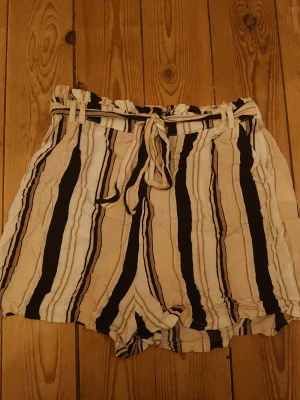 Randiga shorts med knytband - Säljer ett par loose-fit shorts med beige, svarta och vita vertikala ränder. De har hög midja med resår och ett matchande knytband i midjan. Materialet känns lätt och luftigt, perfekt för varma dagar.☺️💕