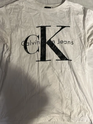 Vit Calvin Klein Jeans t-shirt - Vit t-shirt från Calvin Klein Jeans med stor svart CK-logga och text på bröstet. Klassisk rund halsringning och korta ärmar. Tillverkad i mjuk bomull som är skön mot huden. Perfekt för en clean och stilren look.