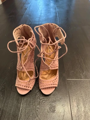 Rosa klackade sandaler från JustFab - Säljer ett par rosa sandaler från JustFab med hög klack och öppen tå. Skorna har snygga utskurna detaljer, snörning framtill och dragkedja bak. Materialet är textil med mockakänsla och de har en feminin, trendig look. Oanvända endast provade 