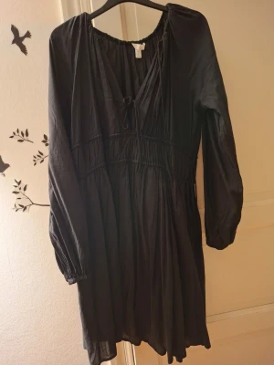 Svart kortklänning med snörning - Säljer en svart kortklänning från H&M med v-ringning och snörning framtill. Klänningen har långa ärmar med resår vid handlederna och ett löst, flowy fall. Perfekt för dig som gillar enkel men snygg stil.