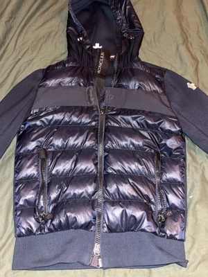 Äkta blå dunjacka från Moncler  - Snygg svart dunjacka från Moncler  med glansig finish, huva och två dragkedjeförsedda fickor framtill. Jackan har quiltad design och ribbade muddar nedtill för extra komfort. Perfekt för lager-på-lager och streetwear-vibe.