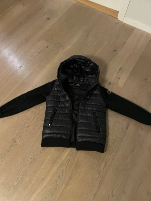 Svart pufferjacka med huva nypris 1600 - Säljer en svart pufferjacka med huva och quiltad kropp. Jackan har släta svarta ärmar i kontrastmaterial och två dragkedjeförsedda fickor framtill. Perfekt för dig som vill ha en stilren och sportig look.