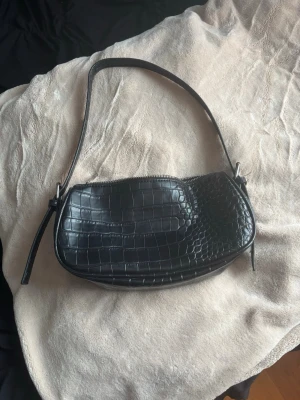 Svart axelväska från NLY Accessories - Säljer en svart axelväska från NLY Accessories i croco-mönstrad syntet. Väskan har en stilren form, dragkedja upptill och ett smidigt innerfack. Perfekt storlek för det viktigaste och enkel att slänga över axeln för en clean look.