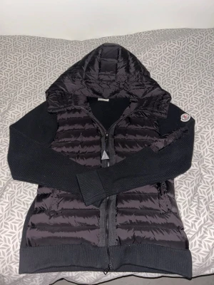 Svart Moncler pufferjacka med huva - Säljer en svart pufferjacka från Moncler med stickade ärmar och huva. Jackan har dragkedja framtill, ribbade muddar och Moncler-logga på ärmen. Perfekt för dig som vill ha en snygg och sportig look med premiumkänsla.