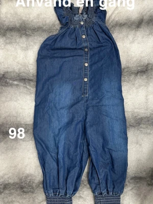 Jeans jumpsuit med knappar, stl 98 - Söt jeans jumpsuit i blå denim med smockdetaljer vid benslut och halsringning. Knappstängning framtill och ärmlös design gör den både bekväm och enkel att ta på. Perfekt för dig som gillar en avslappnad och trendig look.🌸 Stor i storlek. Använd en gång. Storlek 98