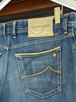Jacob Cohën Limited Edition jeans - Unika blå jeans från Jacob Cohën Limited Edition, nummer 67 av 179. Snygga kontrastsömmar i gult och cool detalj på bakfickan. Tillverkade i japanskt tyg med italienskt hantverk. Klassisk femficksmodell med knappgylf och exklusiva detaljer. Har blivit uppsydda i benen men nu uppsprättade finns en liten tråd som knappt syns. 