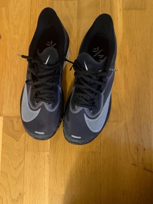 Nike ZoomX svarta löparskor - Säljer ett par svarta Nike ZoomX sneakers med grå detaljer och stor vit swoosh på sidan. Skorna har snörning, ventilerande meshmaterial och en tjock, vit sula för extra komfort. Perfekta för dig som gillar stilrena och sportiga skor.